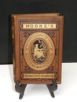 Front cover of Moore's Poetical Works book showing brown cover with intricate black and gold embossed design featuring an oval medallion with a seated figure and floral detailing.