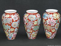 Three vintage Asian porcelain vases, hand decorated with colorful mille fleur floral patterns on a reddish-orange background, shown against a black backdrop.