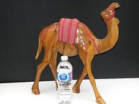 Side view of vintage wooden camel figurine with bottle for size reference showing wood grain and red woven saddle detail.