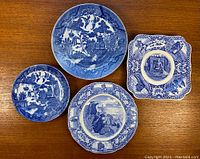 Top-down photo showing four antique porcelain plates with blue and white transferware patterns on a wooden table.