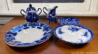 Antique flow blue creamer, sugar bowl, plates, and bowl arranged on wooden surface, showcasing varying blue patterns