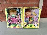 Front view of two Cabbage Patch Kids Babies dolls in original boxed packaging showing dolls and some accessories.