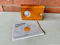 Grundig World Receiver manual lying flat on a gray surface, next to the closed leather cover for the radio, embossed with 'GRUNDIG'