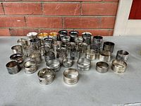 Overview photo showing an assortment of approximately 40 assorted silver plate and pewter napkin rings arranged on a gray surface against a brick wall backdrop.