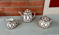 Teapot, cream jug, and sugar bowl arranged on surface showing full set with geometric silver overlay design