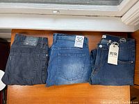 Three pairs of men's pants laid out: Levi's black corduroy pants, Paper Denim & Cloth medium blue jeans with size tag, and Refinery Republic dark blue jeans with tag.