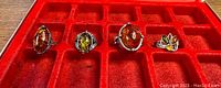 Four sterling silver rings with amber stones displayed on red velvet case. Varying amber colors from deep orange to light yellow-green.