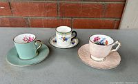 Three vintage Aynsley and Chelson demitasse cups with matching saucers shown together on a grey surface against a brick wall.