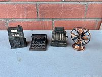 Front view of the four brass colored diecast metal vintage miniatures shaped as slot machine, typewriter, cash register, and fan.