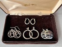Four pairs of circular sterling silver earrings displayed in a brown velvet-lined jewelry box. Designs include small solid hoops, larger rounded hoops, openwork circles with multiple loops, and disc-shaped striped pairs.
