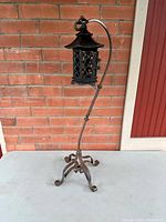 Full view of black wrought iron lantern hanging from a twisted, ornate wrought iron stand with curled feet, placed against a brick wall.