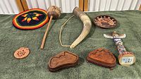 Top down view of all items arranged on green surface showing entire multicultural souvenir collection including hat, carved wooden items, horn flask, painted plate, totem figurine, and carved stone piece.