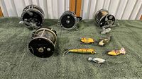 Photo showing five vintage fishing reels placed on green carpet alongside six varied fishing lures