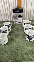 Photo showing six white ceramic mugs arranged around a boxed Maple vacuum coffee carafe on a green towel.