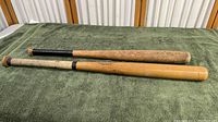 Two vintage Louisville Official Softball bats placed parallel on a green cloth. One bat has black grip tape and worn finish, the other has wrapped handle grip and a lighter wood barrel with printed graphics.
