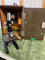 Photo of vintage black metal Technica microscope inside open wooden carrying case resting on green towel.