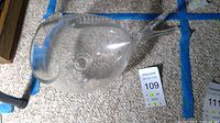 Clear glass fish shaped bowl positioned on carpet next to auction number cards, showing side profile and fin detail.