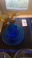 Blue thumbprint glass creamer on matching plate alongside two amber thumbprint goblets on black surface near window.