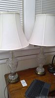 Pair of Hollywood Regency style glass lamps on a wooden table, topped with white fabric shades, showing full height and design.
