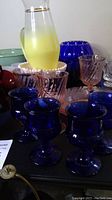 Various vintage glassware including yellow ombre pitcher, pink milkglass bowl, cobalt blue King's Crown goblets, and iridescent glasses.
