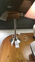 Side view of the silver metal swing arm desk lamp on a wooden surface showing its base, vertical stand, and hinged shade.