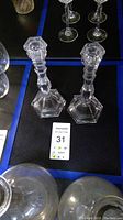 Overall shot of the pair of clear crystal candle sticks with hexagonal bases and faceted stems on display surface with auction lot tag.