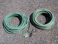 Two coiled green garden hoses on gravel surface, one includes spray nozzle and connector.