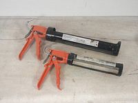 Two caulking guns, one longer and one shorter, showing orange trigger handles and black metal barrels.