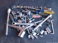 First image showing overall pile of tools including sockets, wrenches, ratchet, drivers, and hex keys arranged on dark surface.