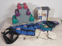 Overview of fitness lot showing dumbbells on rack, resistance bands, weighted straps, fitness DVDs, training gloves, and swim goggles on table