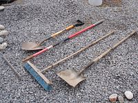 Four garden tools laid on gravel including two shovels, a landscaping rake, and a push broom