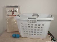 Plastic laundry basket with ventilation holes and two blue dryer balls in front, along with a packaged beige canvas garment storage bag behind them.
