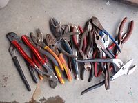 Multiple pliers, channel locks, cutters with varied handle colors and wear, arranged on a concrete floor.