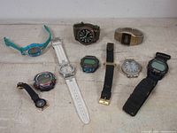 Nine assorted wristwatches laid out on a light surface showing variety of colors, strap materials, and styles