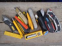 Top view of twelve various utility cutters arranged on wooden surface showing different colors, handle styles and blade types