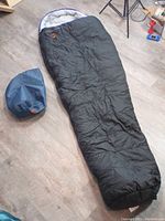 Full view of black Lugger Elite Polarguard sleeping bag laid out on floor with blue storage bag next to it.