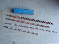 Top-down photo of five masonry drill bits with spiral flutes and rust marks next to a blue plastic case labeled Impact Driver.