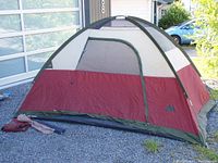 Front and side view of dome tent setup on gravel with poles and stakes visible on ground, color red/beige/green with mesh windows.