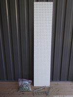 Full view of the white pegboard panel measuring approximately 12 x 60 inches, standing upright against a corrugated metal wall, resting on a wooden floor.