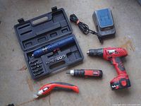 Photo shows a blue cordless screwdriver tool with bits and sockets in black plastic case, a red and black cordless drill with battery, a red and silver cordless screwdriver, a red utility knife, and a black battery charger with cord.