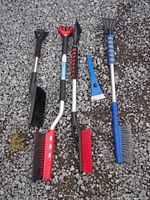 Five snow brushes and ice scrapers displayed on gravel surface showing various handle styles, bristle colors, and sizes.
