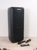 Front view of black rectangular Sylvania SP961 karaoke speaker with microphone, remote control, and cable arranged in front on white background.