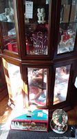 Full view of glass display cabinet with porcelain snow babies and assorted Christmas decorations, box of glassware, and snow globe on floor.