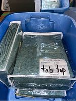 Blue plastic bin filled with textured green fabric tablecloths in original packaging marked 'tabletop' with size 42" x 95"