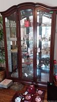 Full front view of the wood framed display cabinet showing three glass doors with decorated glass and contents inside, five glass shelves, and a reflected light source.