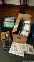 Box with multiple brand new 11x14 and 10x12 size photo frames and smaller boxes of silver plated frames plus white frames and prints on hardwood floor.
