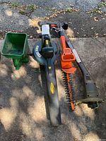 Four gardening tools: green Yardworks electric leaf blower with black nozzle, orange Black & Decker hedge trimmer, black Black & Decker weed whacker, green Scotts seed spreader.