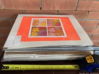 Stack of sealed art prints with floral design on top and measuring tape showing size approximately 14 inches wide