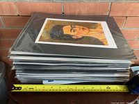 Stack of 26 matted art prints in sealed plastic sleeves, resting on a surface with a measuring tape showing approximately 17 inches width.
