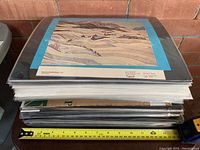 Stack of sealed and matted prints with a winter landscape by Grandma Moses on top, ruler included for size reference